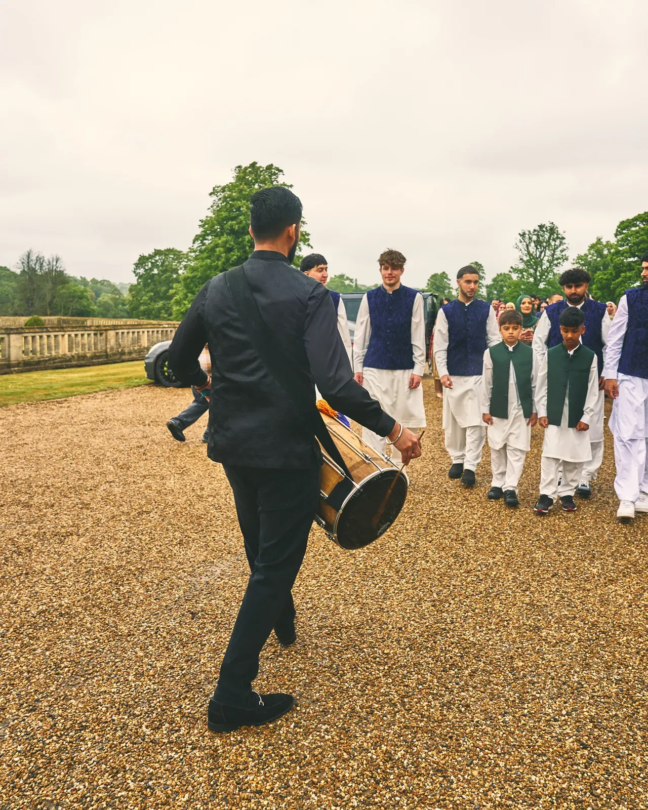 Dhol player leading the groom's party up a gravel drive