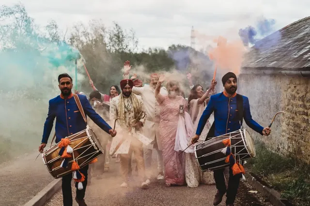 Two dhol players leading a baraat through coloured smoke