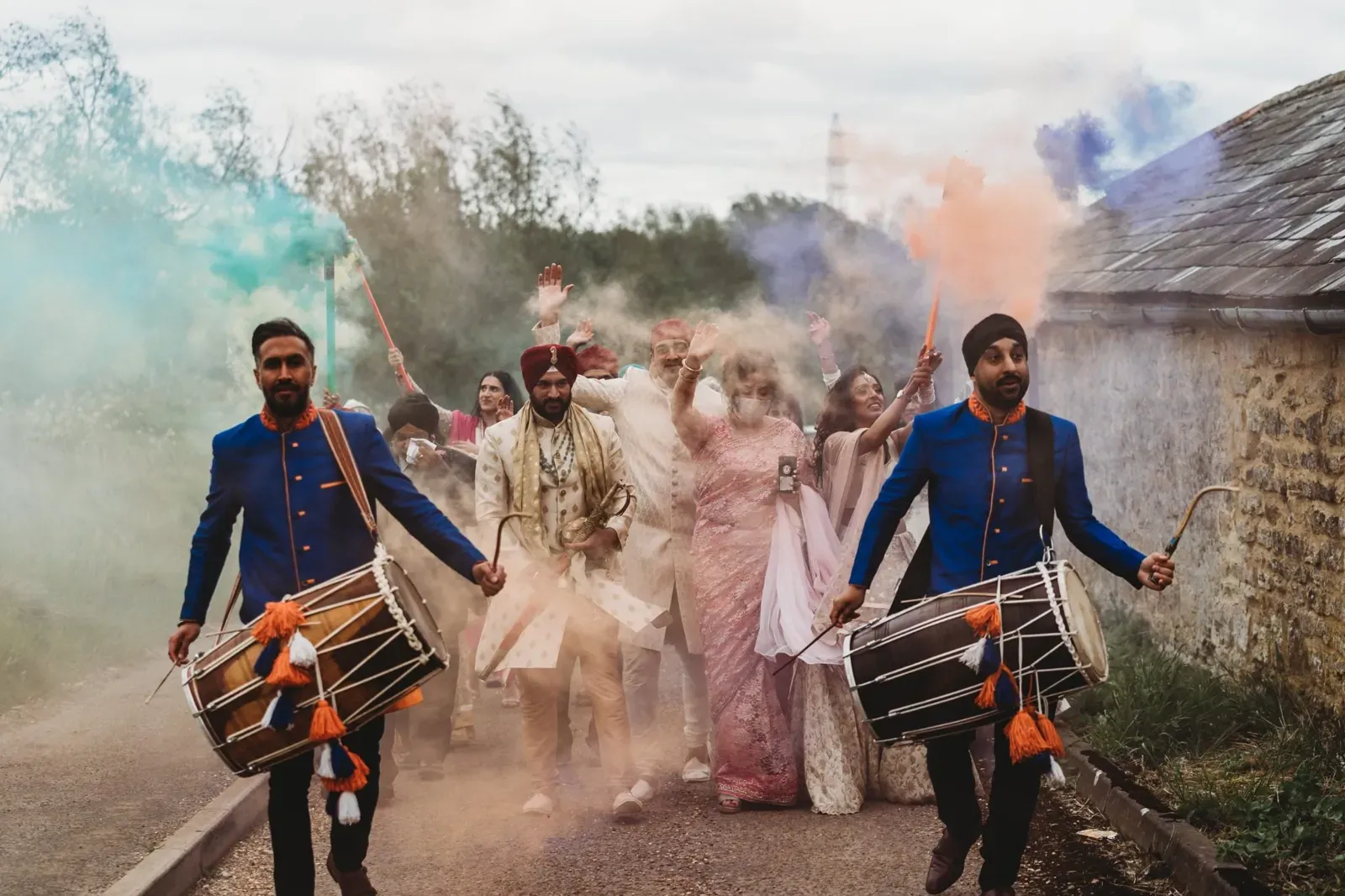 Two dhol players leading a baraat through coloured smoke