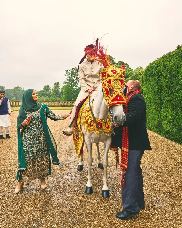 Groom on a white horse in red and gold baraat costume, welcomed by family