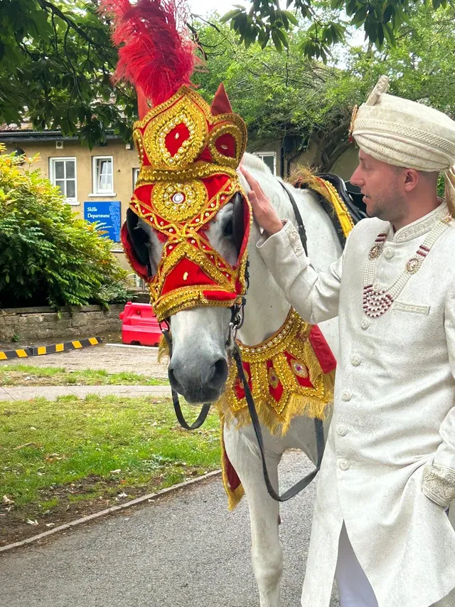 Groom in a cream sherwani beside a white horse in a red and gold headdress